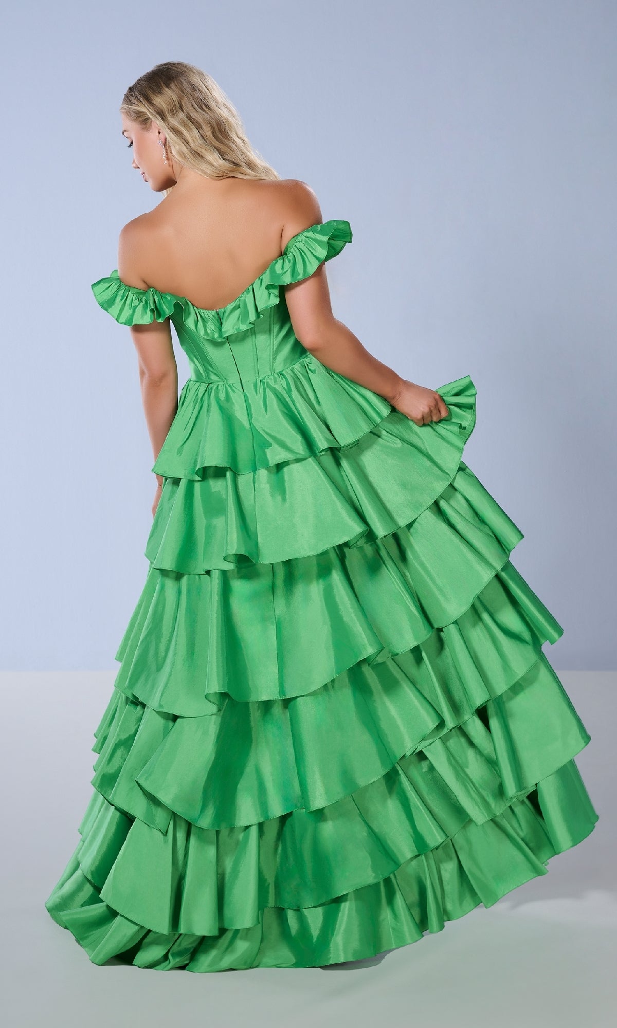 A woman faces away in the Tiffany 16175 long prom dress, featuring a bright green off-the-shoulder corset bodice and layered ruffled tiers, set against a plain light background.